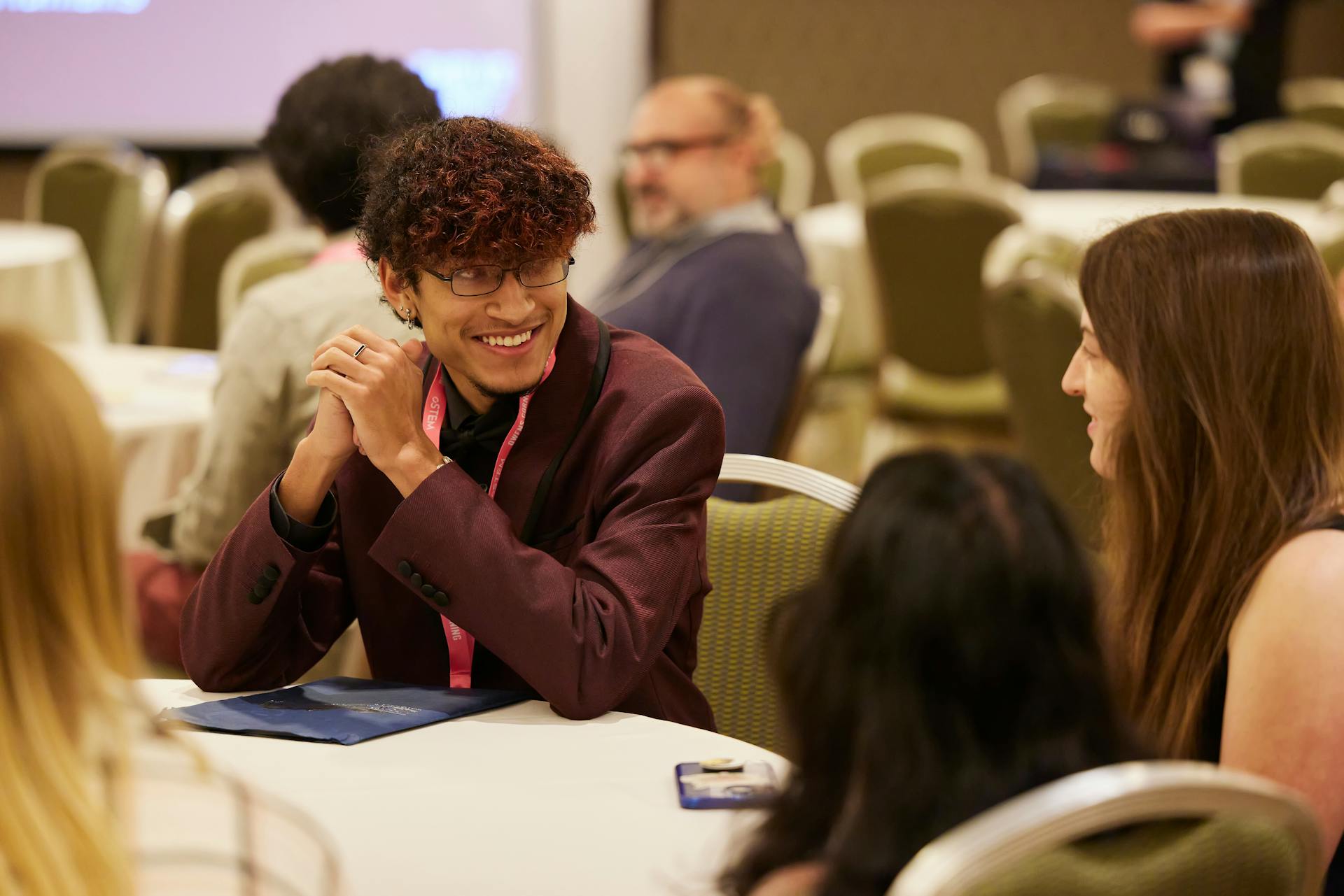 A decorative image of oSTEM conference attendees sitting around a table. One attendee is in focus looking at another attendee, while four other attendees can be seen blurred in the foreground and background.